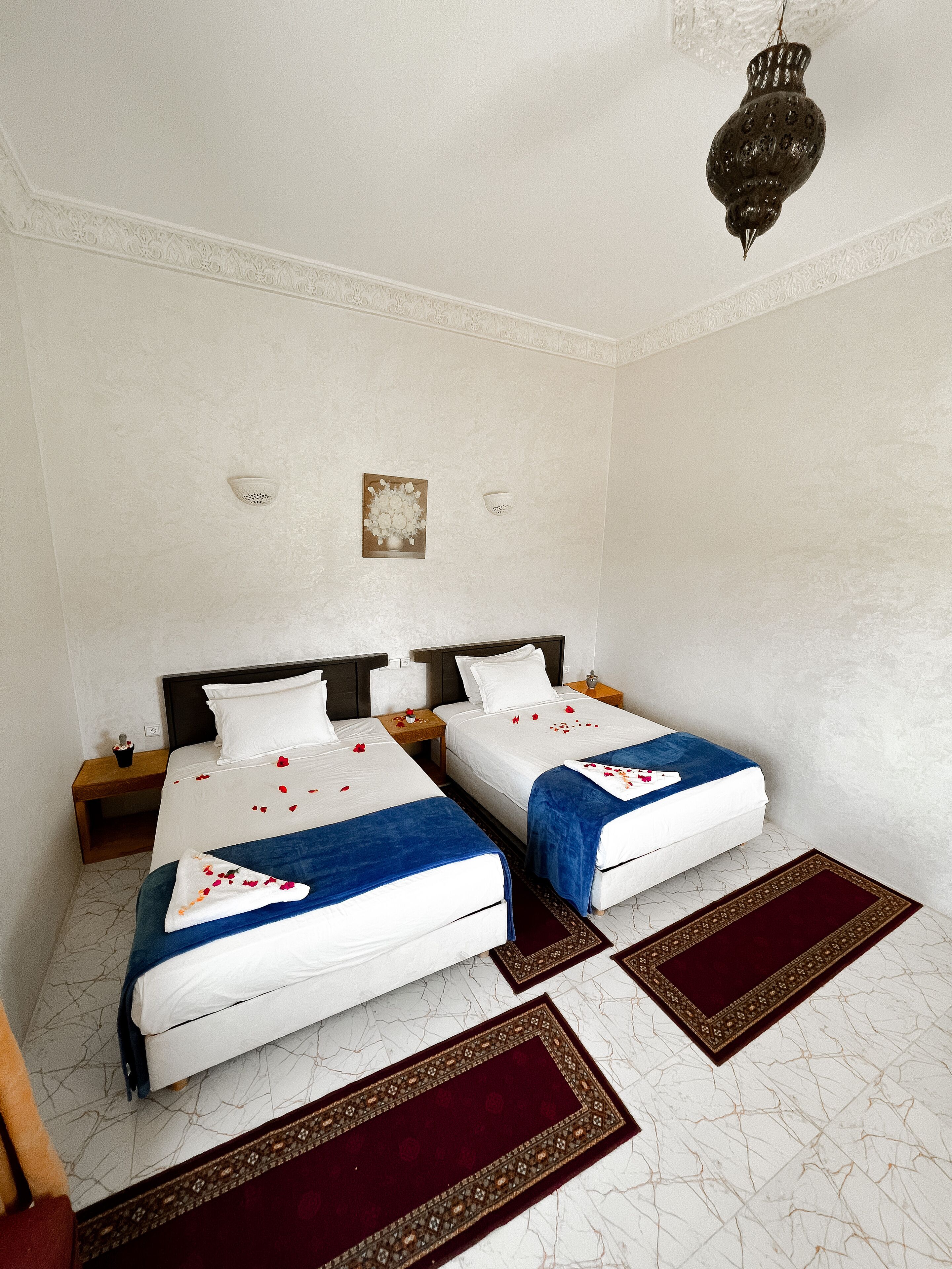 A hotel room with two beds, white walls, decorative ceiling medallions, a chandelier, and a small wall-mounted artwork.