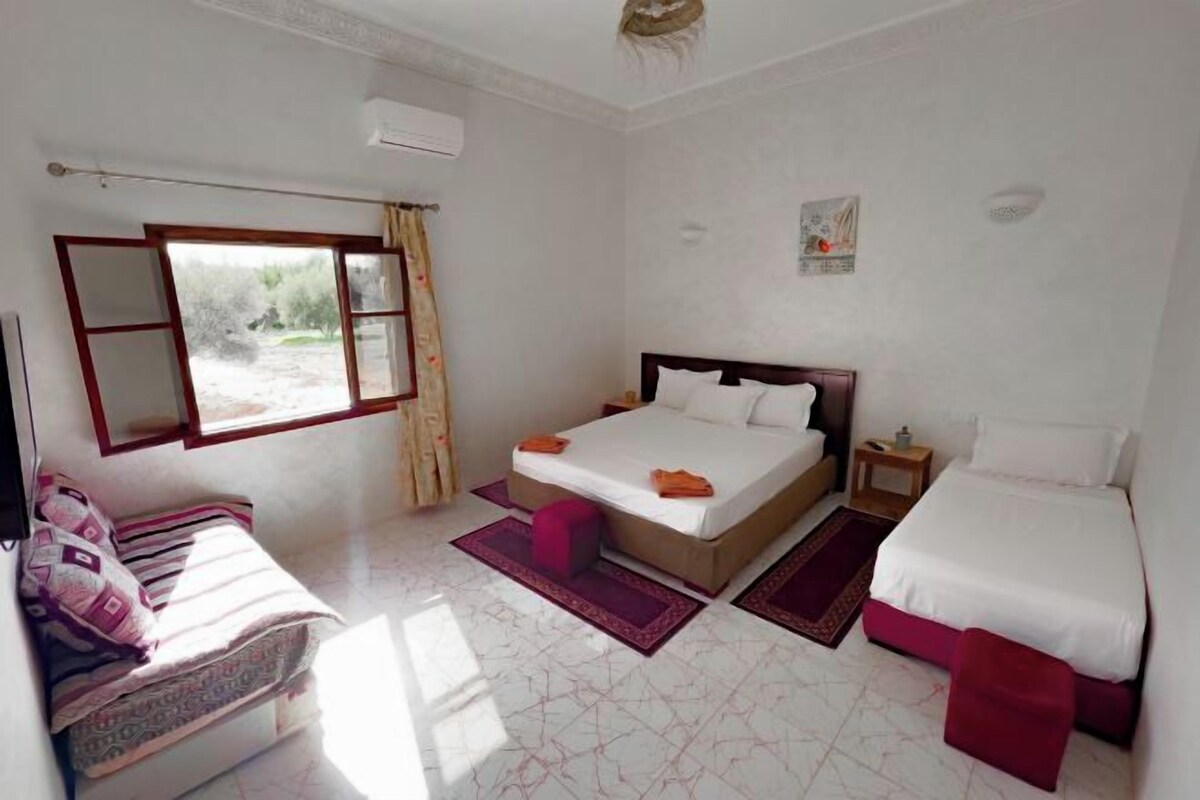 A bedroom with a large bed, a small bedside table, a chair, and a window.