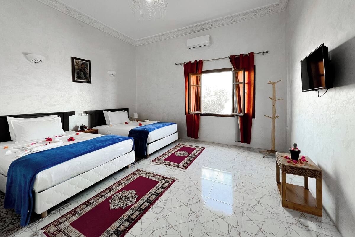 Une chambre d’hôtel avec deux lits, une télévision, une fenêtre avec des rideaux, un ventilateur de plafond et une petite table sur laquelle est posé un vase avec des fleurs.