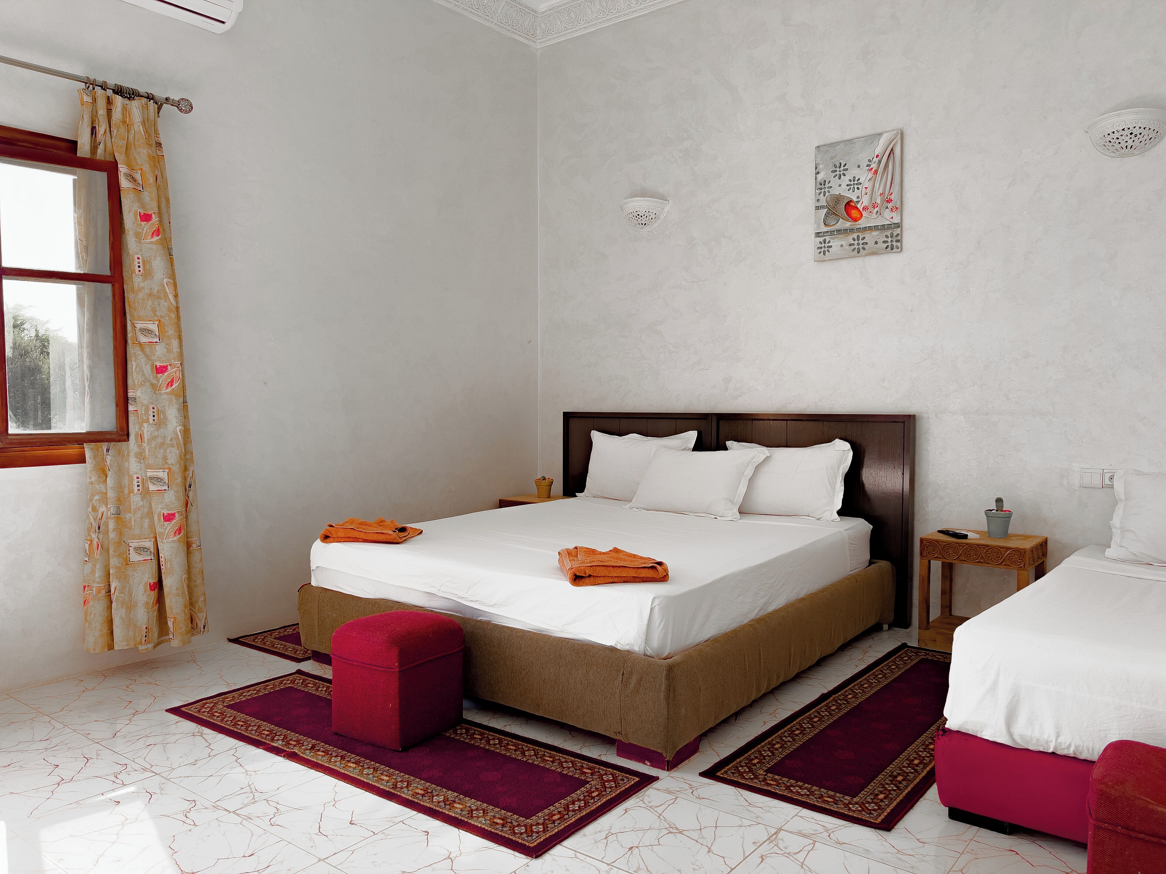 Un dormitorio ordenado con una cama, una mesita de noche, un otoman rojo y una ventana con cortinas.