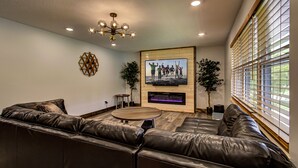 A modern living room with a leather sofa, a wooden coffee table, a fireplace, and a large window with blinds. - Modern! 4BR, Game Room, Pets, Hot Tub, PS5, 6 min to Dollywood + More! Sleeps 8! (Pigeon Forge)