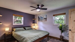 A bedroom with a bed, a ceiling fan, two windows, a nightstand, a bedside lamp, and a potted plant. - Modern! 4BR, Game Room, Pets, Hot Tub, PS5, 6 min to Dollywood + More! Sleeps 8! (Pigeon Forge)