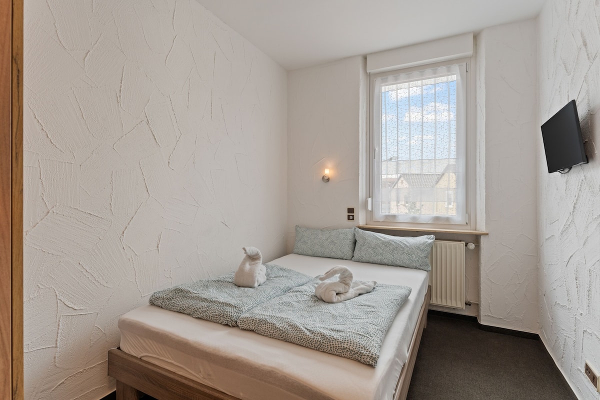 A bedroom with a bed, two pillows, a window, a wall-mounted TV, and a radiator.