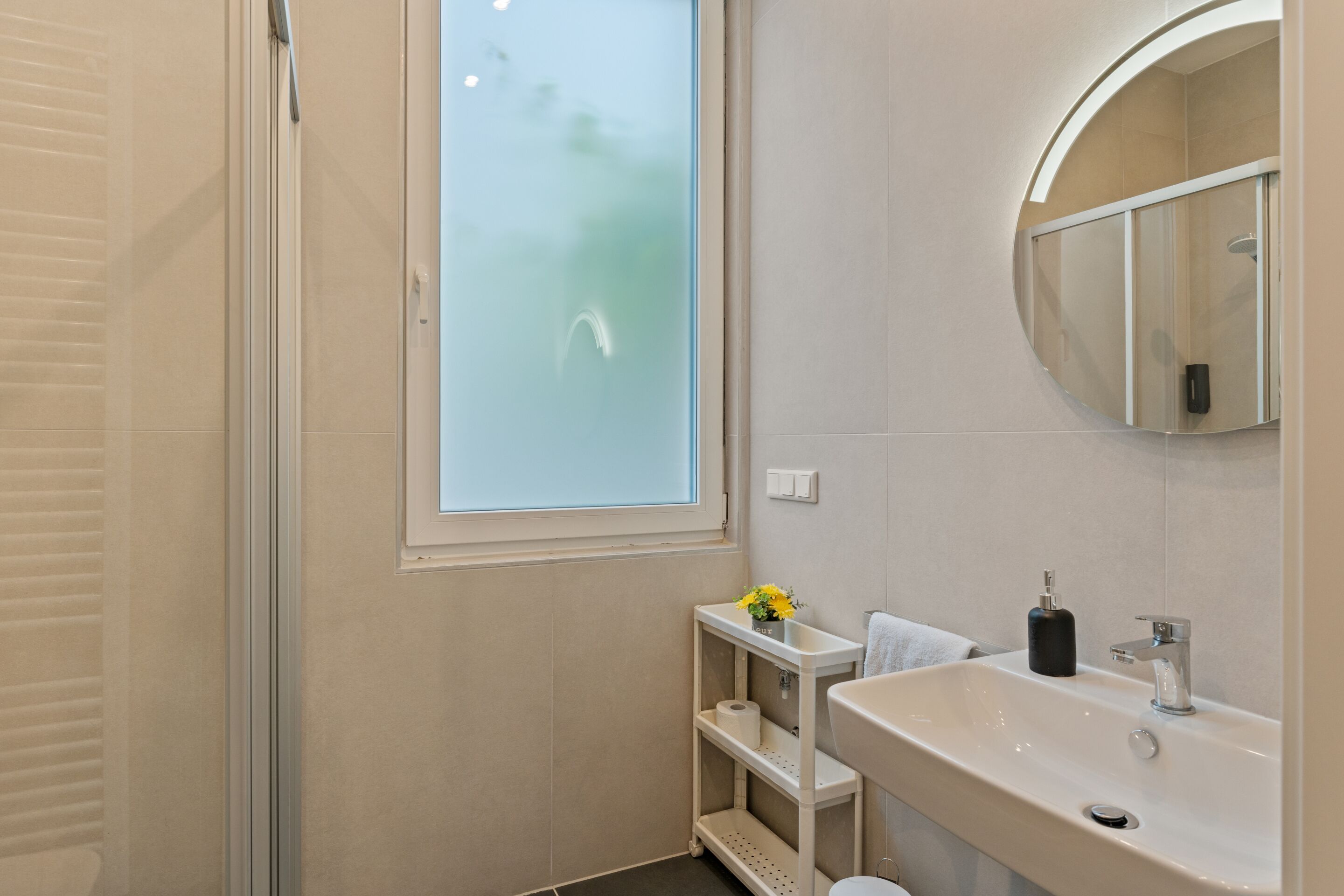 A bathroom with a glass shower door, a sink with a mirror, and a shelf with toiletries.