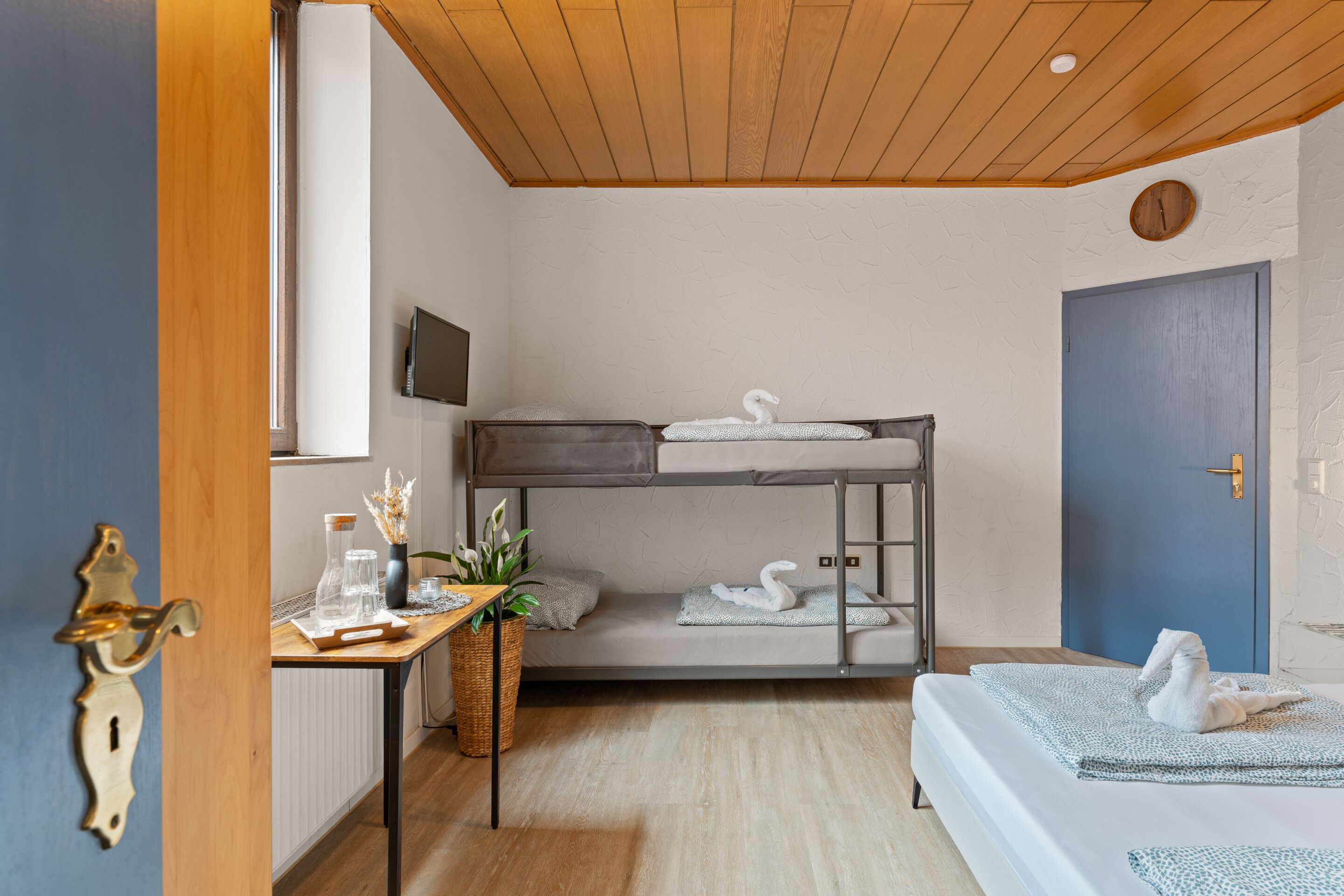 Une chambre avec un lit superposé, un plafond en bois, une petite table avec une plante et une porte bleue.