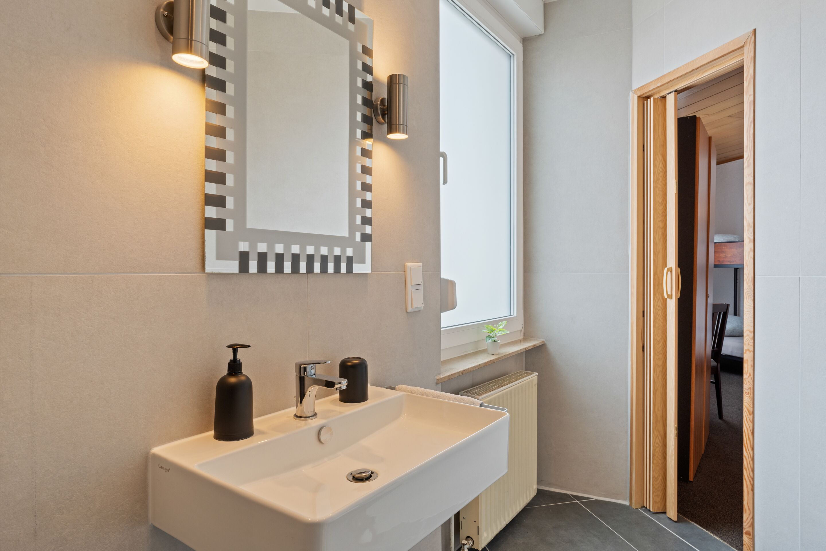 Une salle de bain moderne avec un grand miroir, un lavabo blanc et une fenêtre laissant entrer la lumière naturelle.