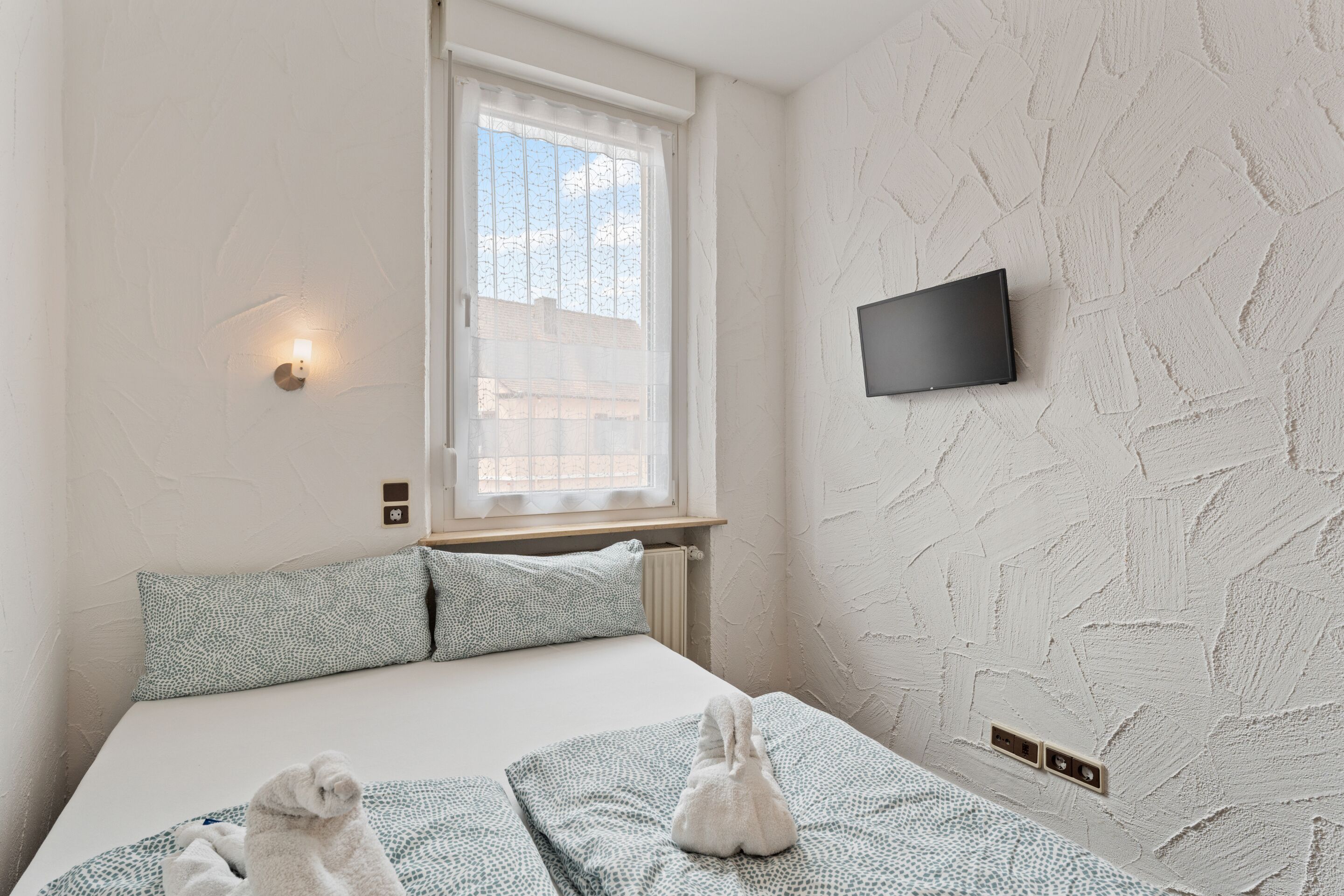 A bedroom with a bed, pillows, a window, a wall-mounted TV, and a wall with a textured pattern.