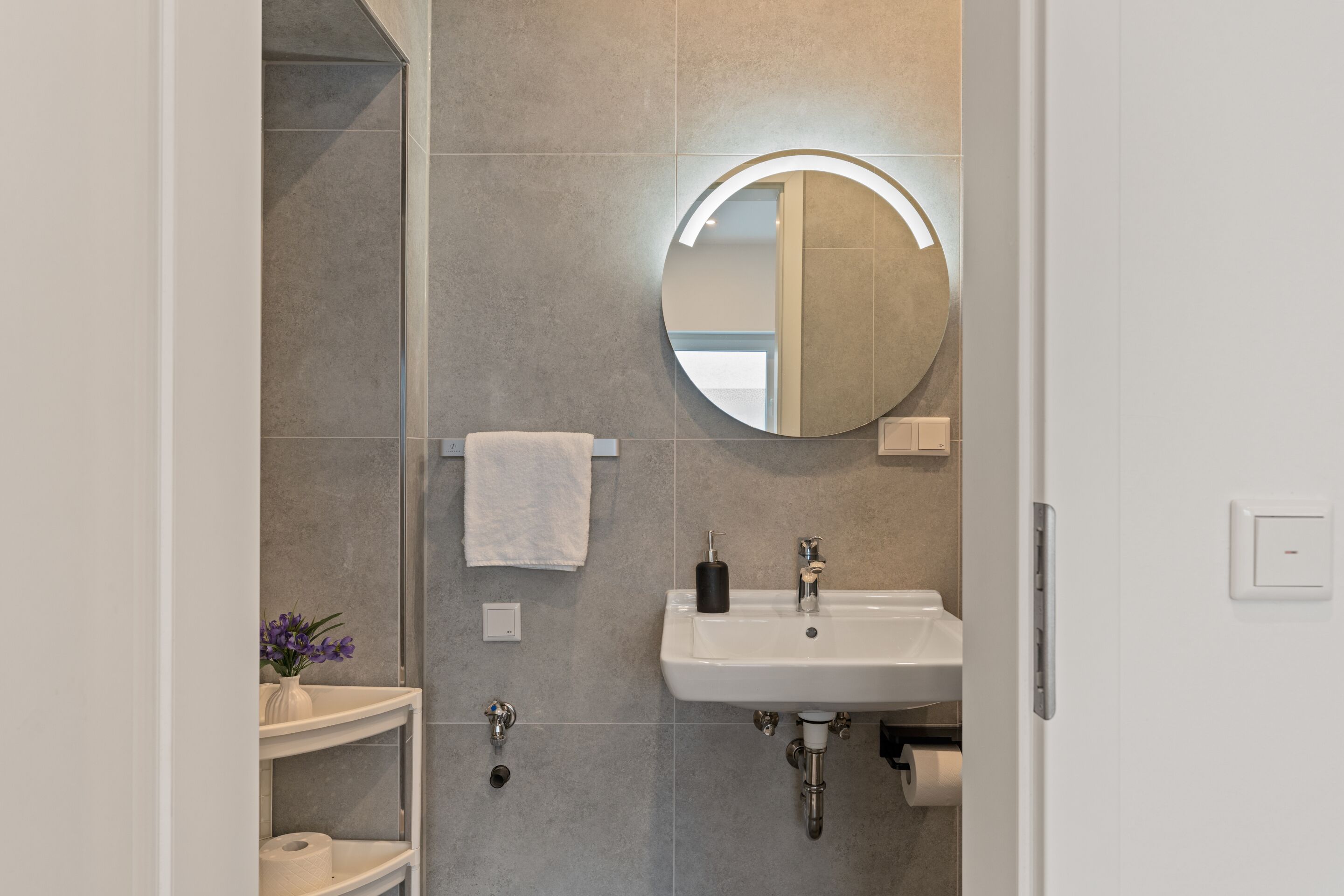 Ein modernes Badezimmer mit einem weißen Waschbecken, einem runden Spiegel und einem Handtuch an einem Halter.