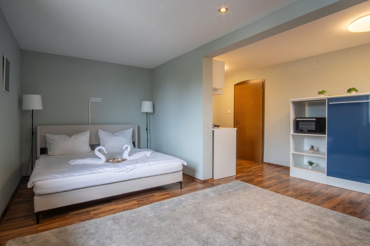 A bedroom with a bed, bedside lamps, a wooden floor, a white wardrobe, and a door leading to another room.