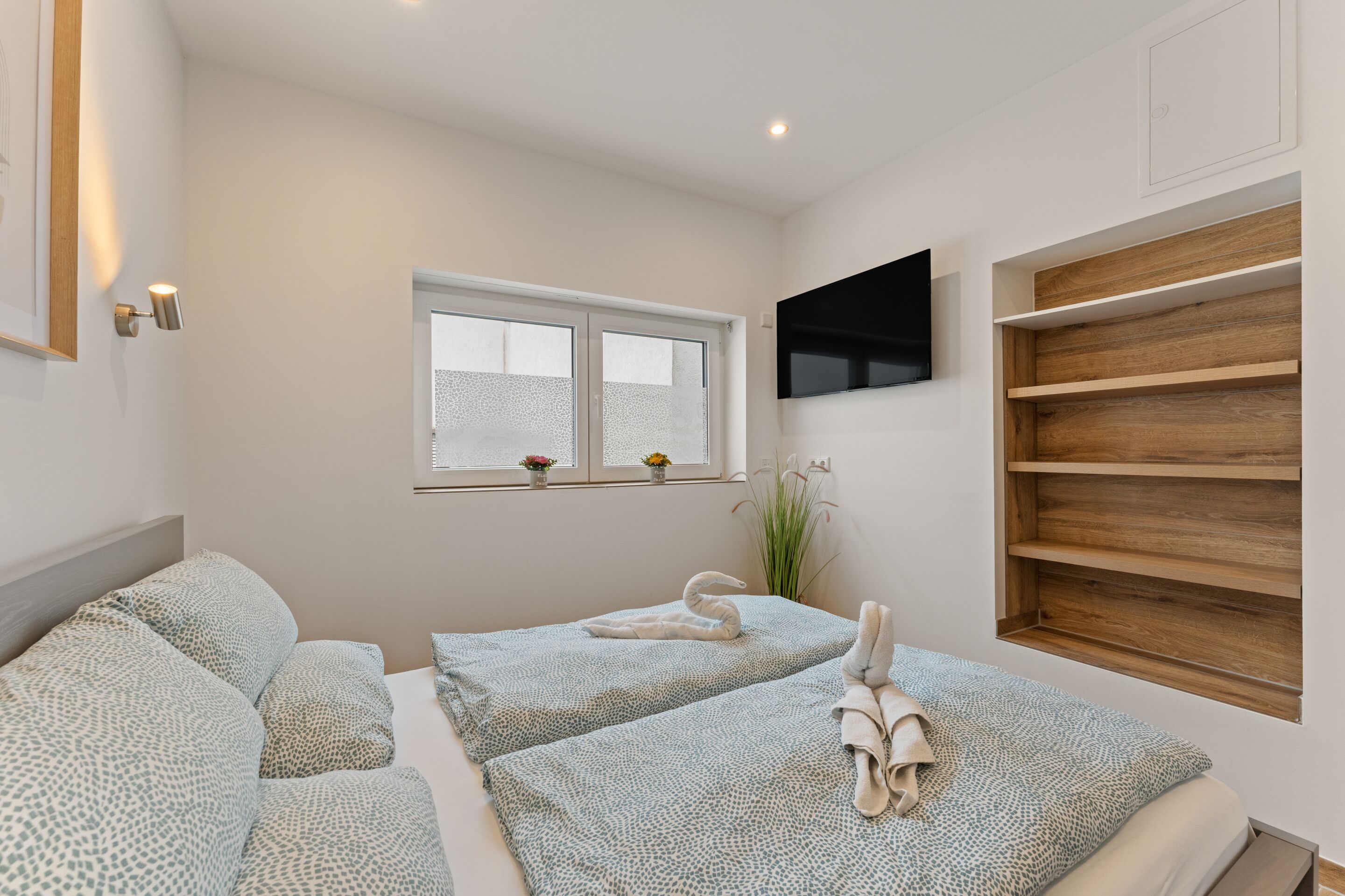 A bedroom with two beds, a TV mounted on the wall, a window with a view, and a wooden shelf unit.