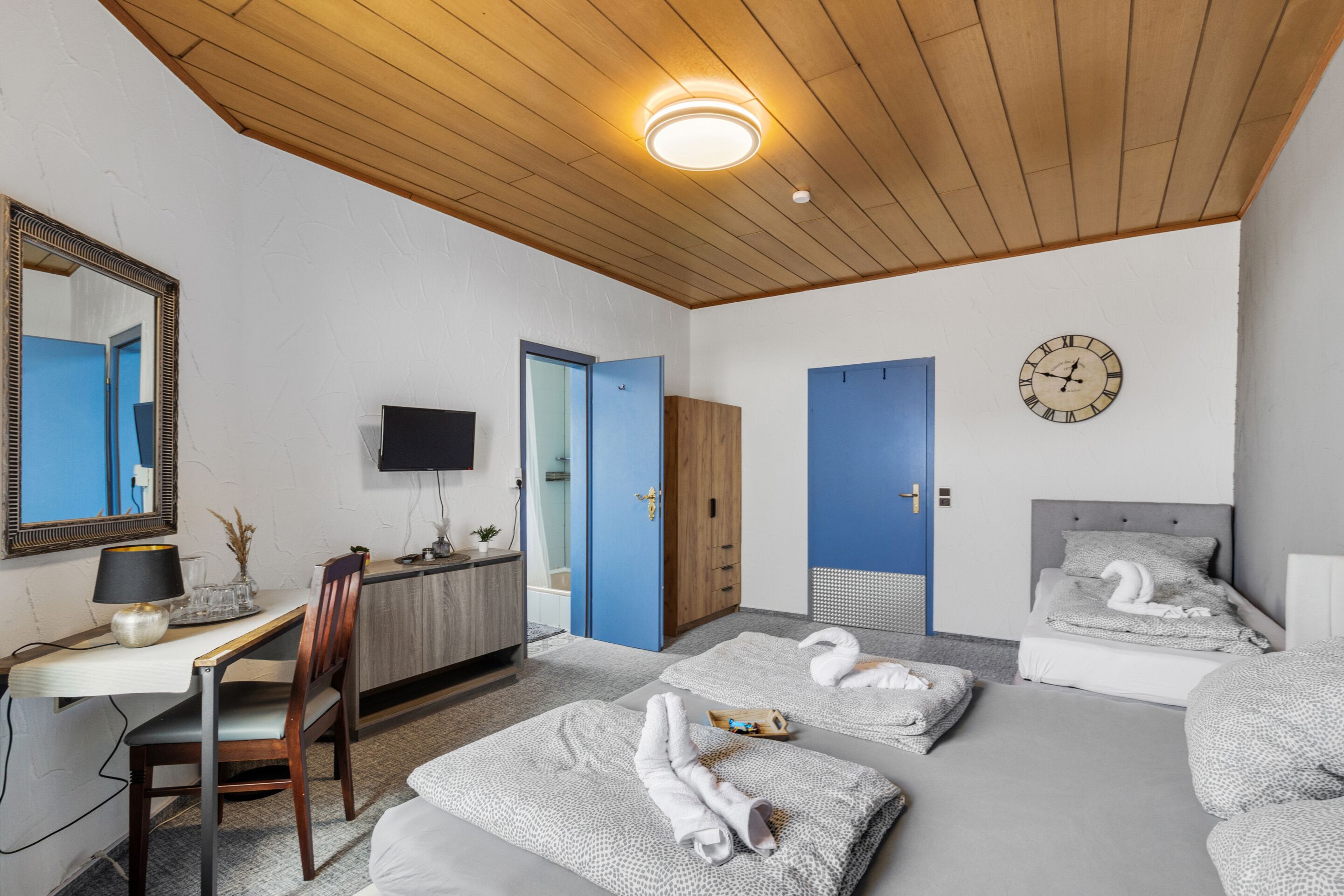 Een hotelkamer met een houten plafond, een bureau met stoel, een televisie, een bed met twee kussens, en een klok aan de muur.