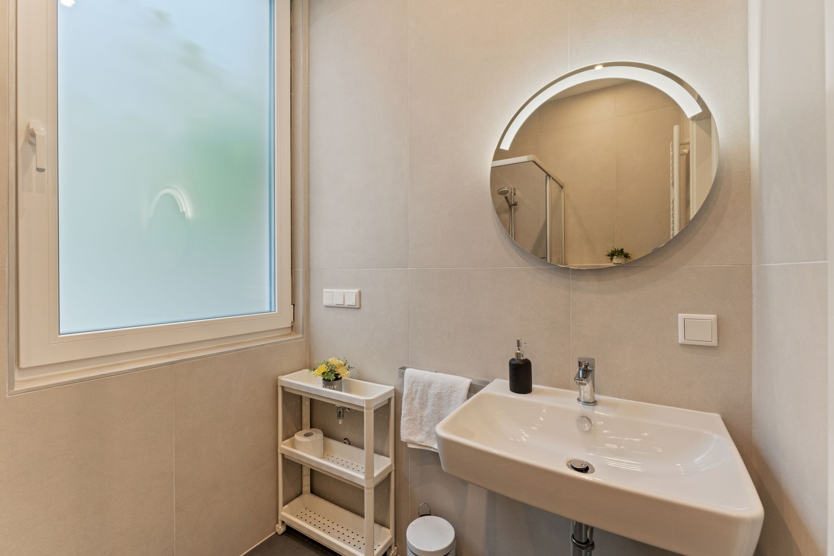 A bathroom with a white sink, a round mirror, a small shelf with toiletries, and a window with frosted glass.
