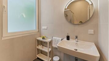 A bathroom with a white sink, a round mirror, a small shelf with toiletries, and a window with frosted glass.
