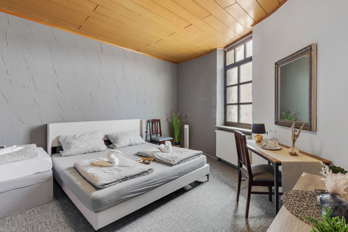A modern bedroom with a large bed, a desk, and a chair. There is a window providing natural light and a mirror on the wall.