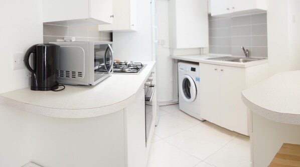 Modern kitchen with stainless steel range hood, microwave, gas stove, and washing machine, set against a backdrop of white cabinetry and a tiled floor.
