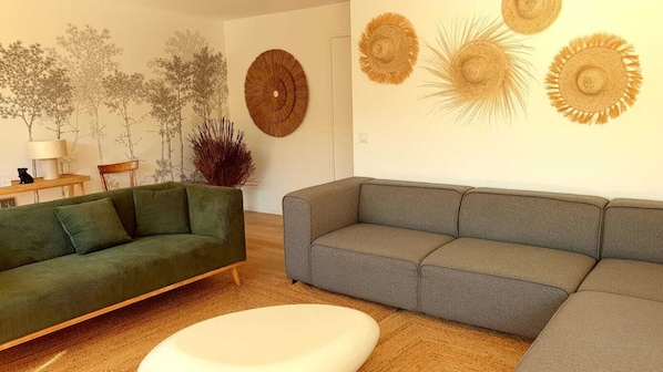 Uma sala de estar com um sofá verde, um sofá cinza e um banquinho branco. Há quadros nas paredes e uma planta.