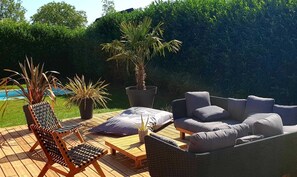 An outdoor patio with a wicker sofa, a wooden chair, and a small table. There is a potted plant and a large planter with a palm tree. - Maison au Calme Avec Piscine (Branville)