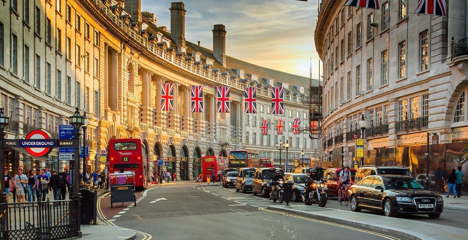 A busy street with double-decker buses, cars, and motorcycles. There are buildings with British flags and a sign for the Underground.
