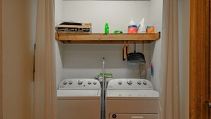 Une buanderie équipée de deux machines à laver et deux sèche-linge superposés, d’une étagère en bois où sont rangés les produits de nettoyage, et d’un balai suspendu.