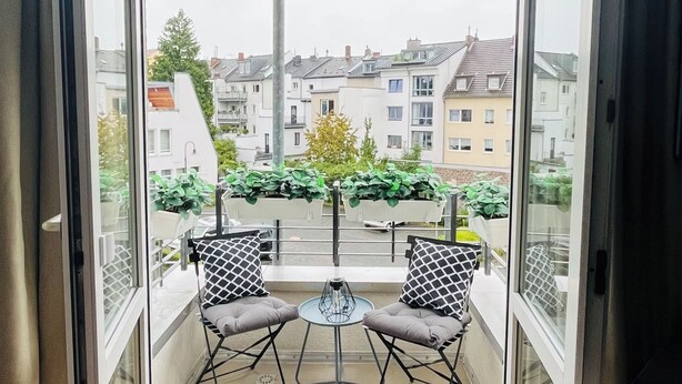 Un balcon avec deux chaises et une petite table, donnant sur un quartier résidentiel avec des maisons et de la verdure.