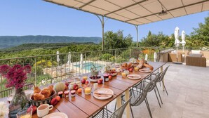 Mesa de desayuno dispuesta con cruasanes, frutas y bebidas, con vista a una piscina y un paisaje exuberante.