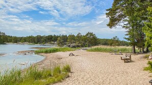A sandy beach with picnic tables and a bench, surrounded by trees and a body of water.
