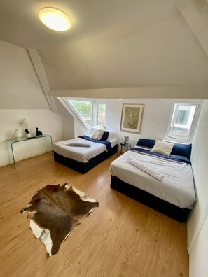 Two single beds with white linens and dark blue accents, a glass coffee table, and a framed botanical illustration adorn this cozy attic retreat.