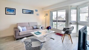 A modern living room with a sofa, coffee table, and two chairs. There are two framed pictures on the wall, a large window with a view, and a flat-screen TV. - A Deux Pas du Port - Vannes (VANNES)