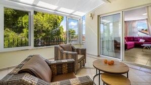 A sunlit room with wicker furniture, a glass door leading to a balcony, and a view of trees and a building. - 4 bedroom amazing home in Zaboric (Zaboric)