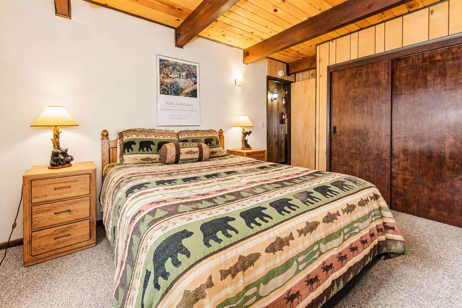Un dormitorio con cama, cabecera de madera, dos mesitas de noche con lámparas, un cuadro enmarcado en la pared y un armario de madera.