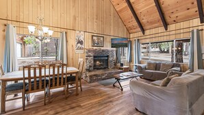Una habitación con paneles de madera, chimenea, una mesa de comedor con sillas, un sofá y ventanas amplias.