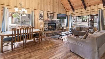 A wooden-paneled room with a fireplace, a dining table with chairs, a sofa, and large windows.