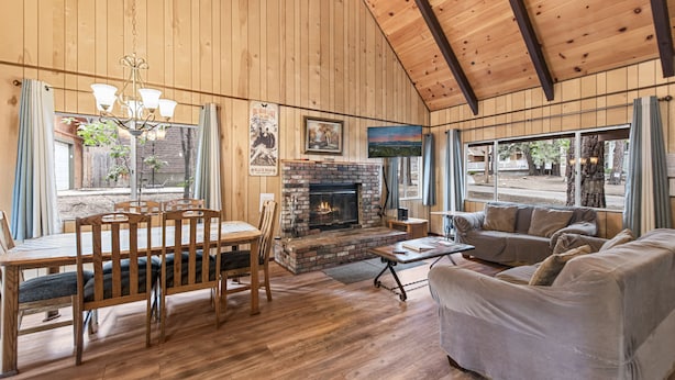 A wooden-paneled room with a fireplace, a dining table with chairs, a sofa, and large windows. - Bear Crossing (Big Bear Lake)