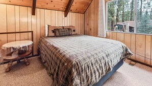 Un dormitorio de cabaña de madera con una cama, una pequeña mesa redonda y una ventana con vistas a árboles.