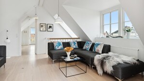 En modern lägenhet i loftstil med högt i tak, en mörkgrå hörnsoffa med färgglada kuddar, ett svart soffbord med en vas och böcker, och ett stort fönster med utsikt över staden.