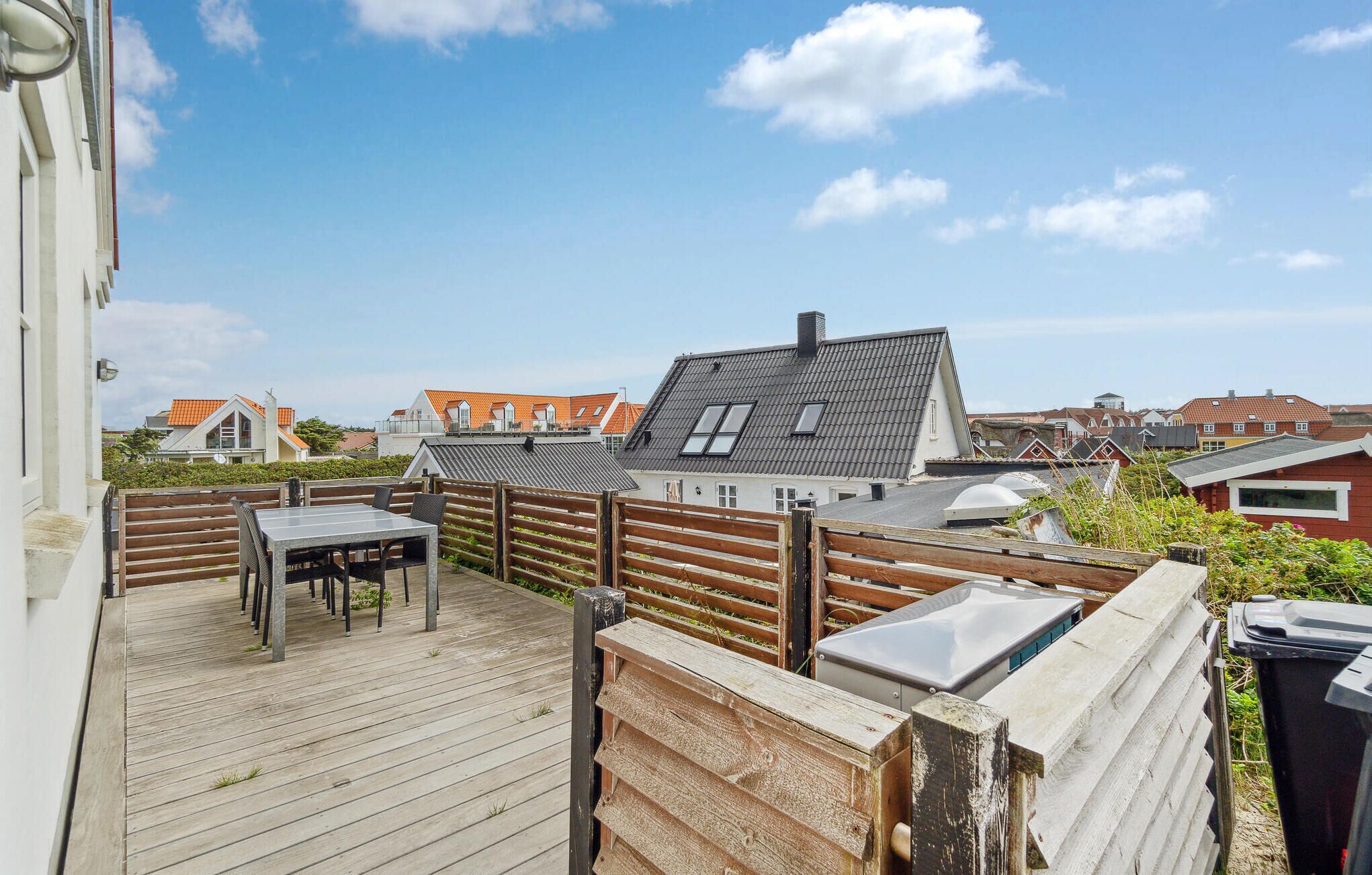 A wooden deck with a table and chairs, surrounded by a wooden fence, overlooking a residential area with houses.