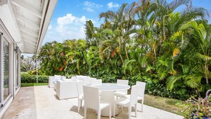 Eine Terrasse mit weißen Korbmöbeln und einem Glastisch, umgeben von üppigen tropischen Pflanzen.