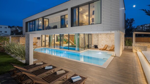 Ett modernt hus med en stor swimmingpool, ett trädäck och rum som är inramade av glasväggar.