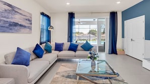A modern living room with a beige sofa, blue cushions, a glass coffee table, and a view of a balcony.