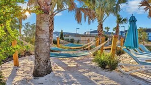 A sandy beach area with hammocks and lounge chairs, surrounded by palm trees and a clear sky.
