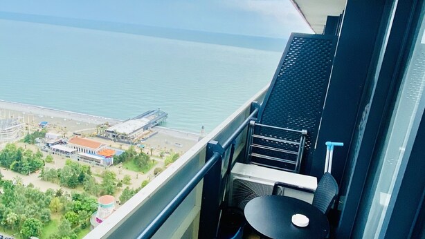 Een balkon met uitzicht op zee, een kleine tafel met een stoel, en een raam met luxaflex.