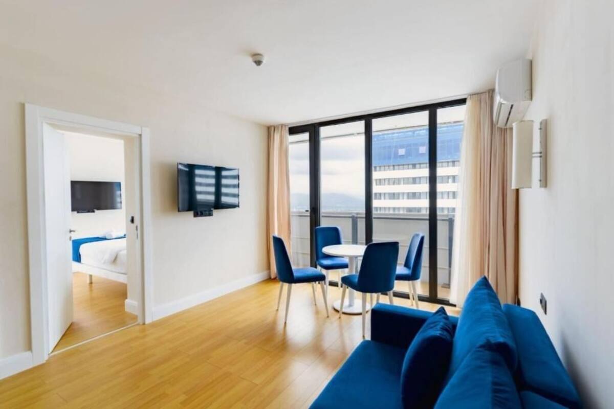 Een moderne hotelkamer met houten vloer, een blauwe bank, een zithoek met stoelen, een aan de muur bevestigde televisie en een groot raam met uitzicht op de stad.