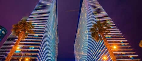 Two tall, modern skyscrapers with a mirrored facade, surrounded by palm trees and illuminated by street lights.