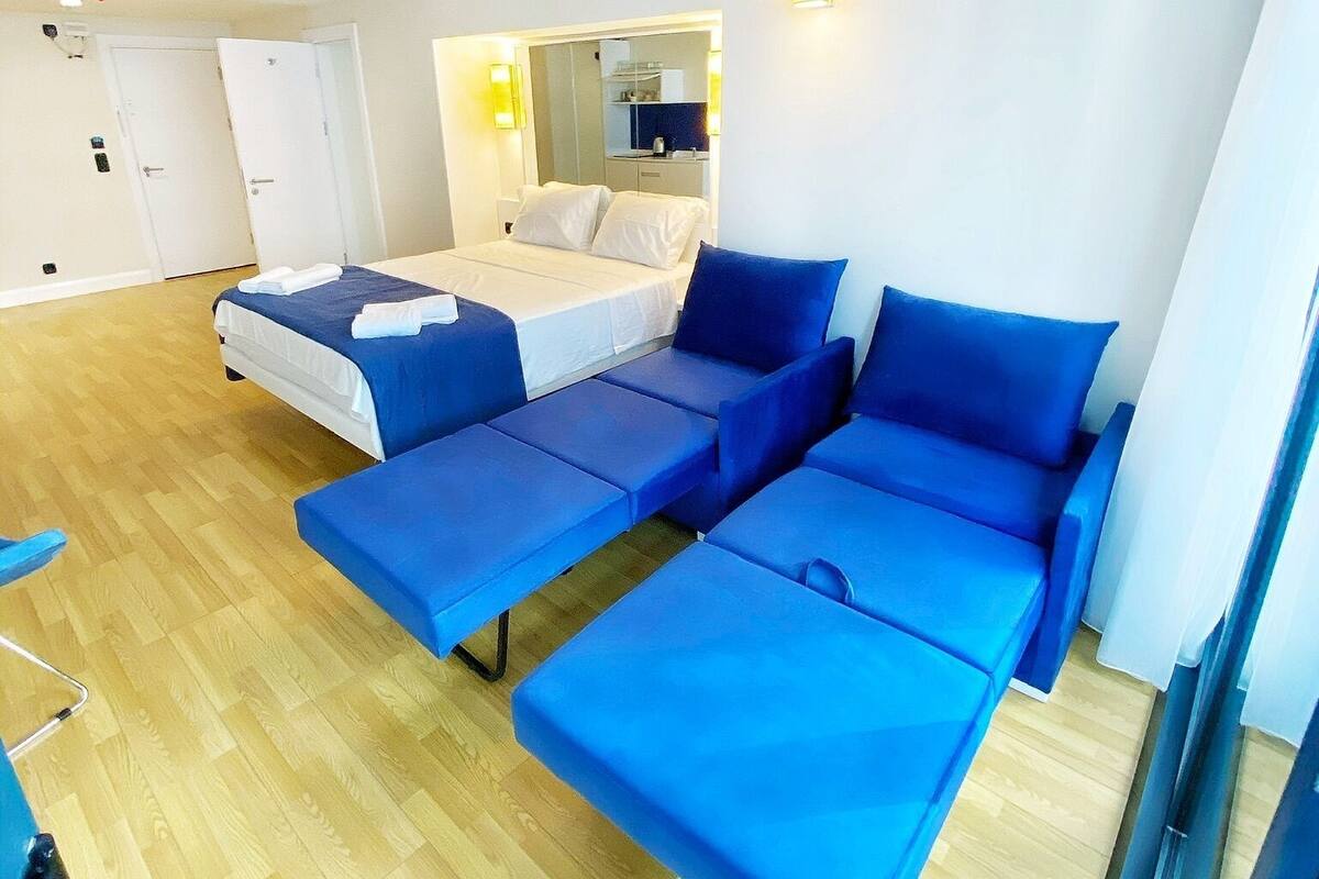 Een moderne hotelkamer met een groot bed, een slaapbank en een kleine tafel. Er is een spiegel boven de slaapbank en een raam met gordijnen.