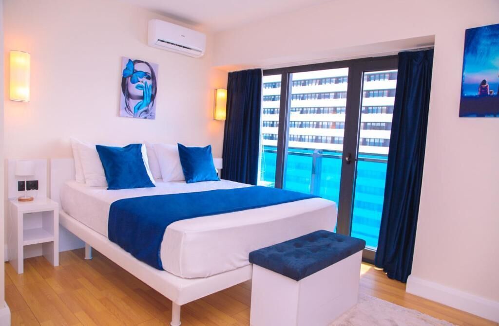 A modern hotel room with a large bed, a small bedside table, a bench, and a balcony with a view of a building and a pool.