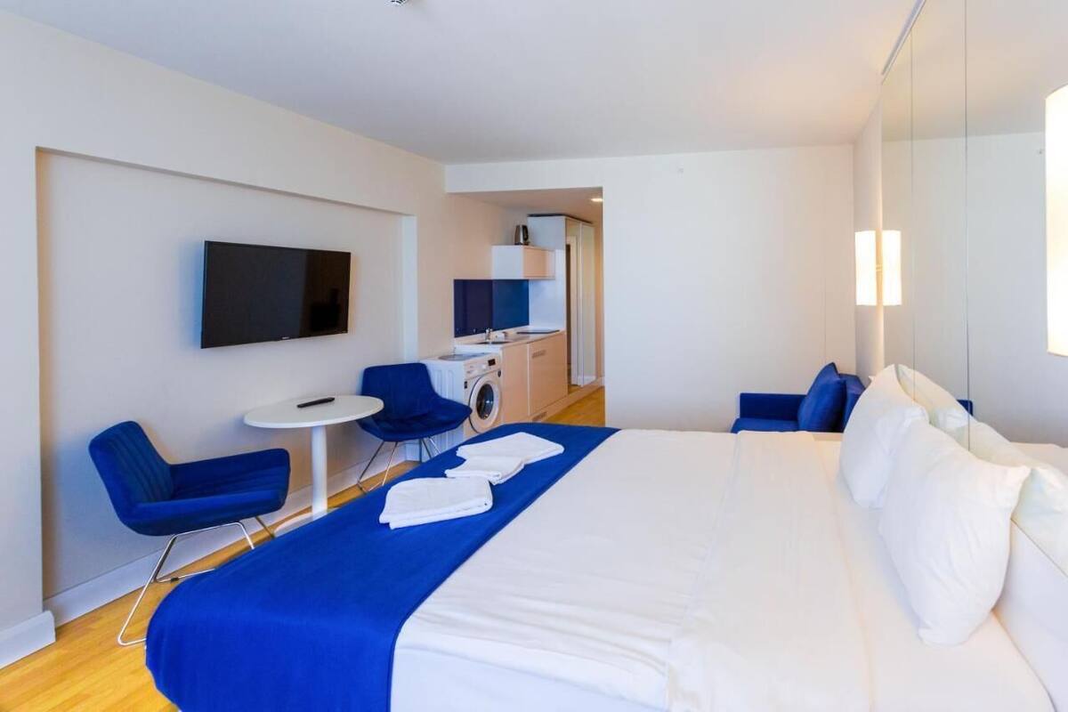 Een moderne hotelkamer met een groot bed, een flatscreen televisie aan de muur, een klein rond tafeltje en twee blauwe stoelen.