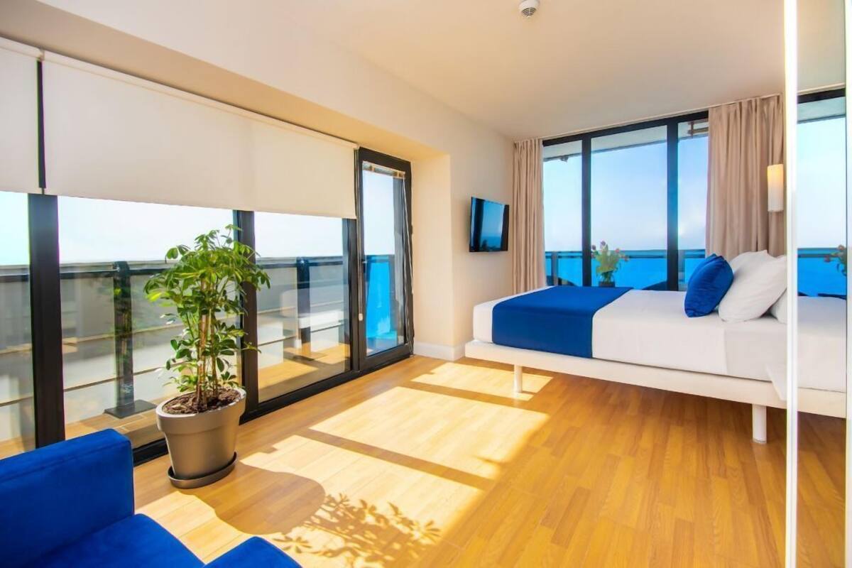 Een moderne hotelkamer met balkon, een grote potplant, een witte bank met blauwe kussens en een flatscreen televisie.