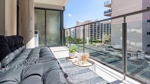 Een balkon met een grijze bank, een klein tafeltje met drankjes en een uitzicht op omliggende gebouwen.