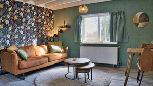 En hyggelig stue med en brun lædersofa, to runde træborde, en blød måtte, en radiator og et vindue med gardiner.
