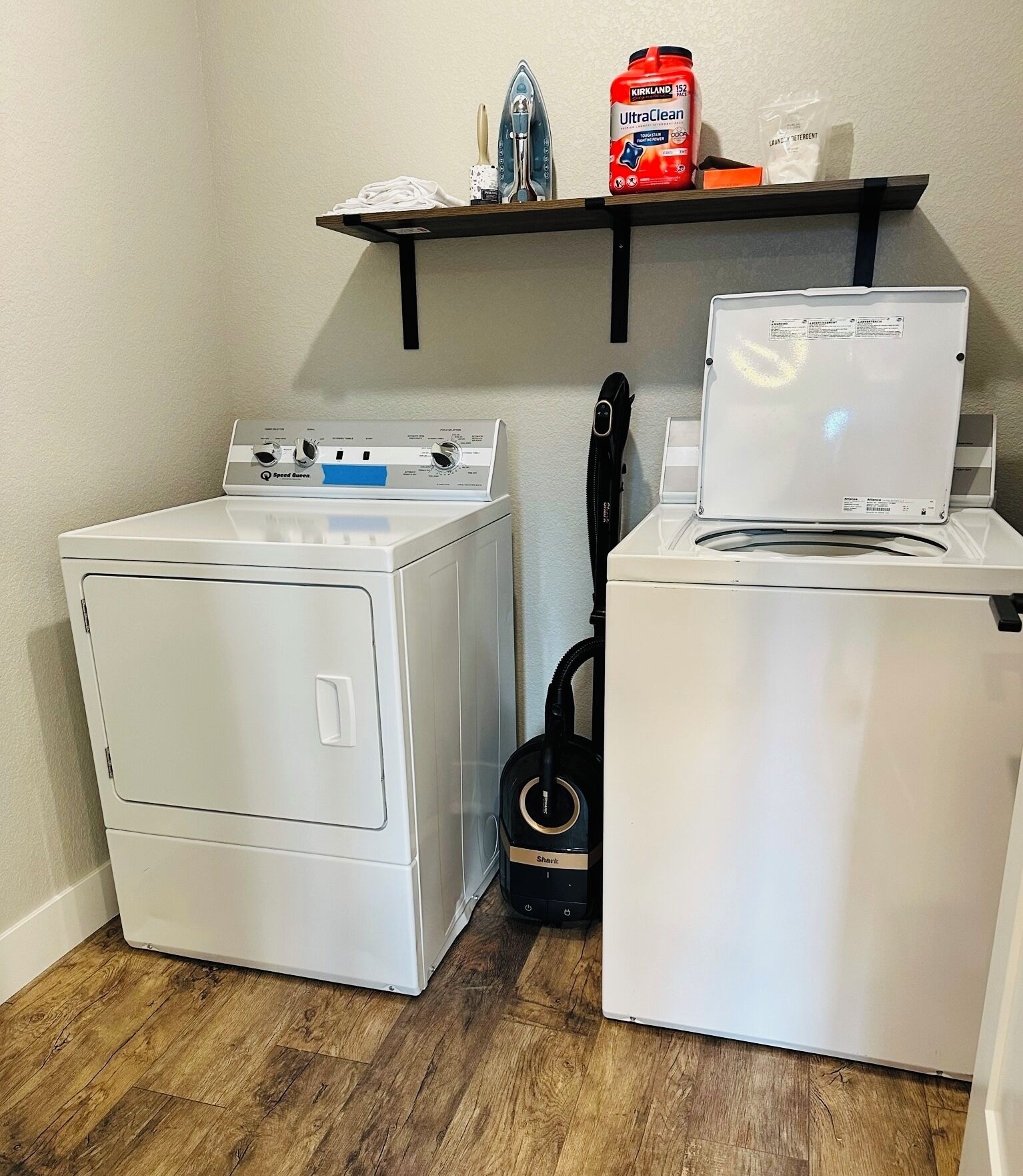 A laundry area with two white washing machines, a shelf with cleaning supplies, and a vacuum cleaner.